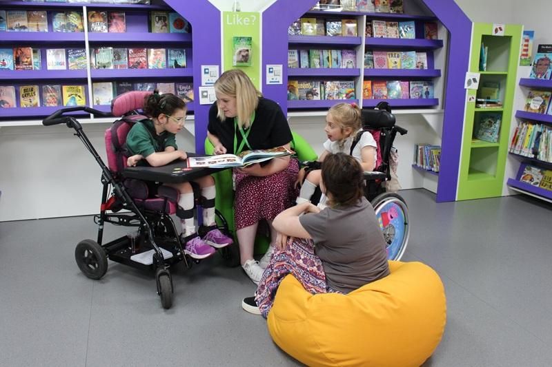 Teacher reading a book to two Treloar's primary students and a support assistant sitting on a yellow beanbag in a colourful library.