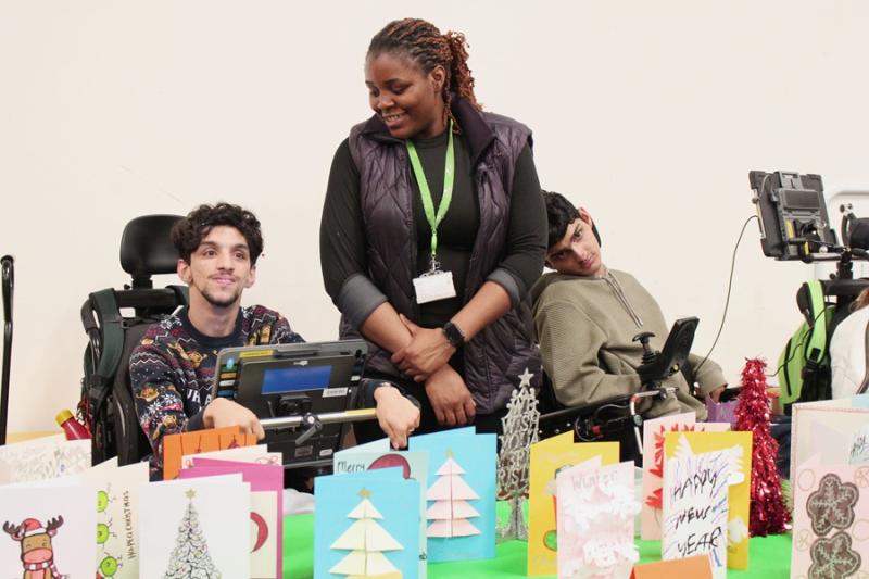 Two Treloar College students and their assistant standing between them at a table displaying colourful handmade holiday cards and small festive decorations. The boys are patiently waiting for customers to arrive at their stall.