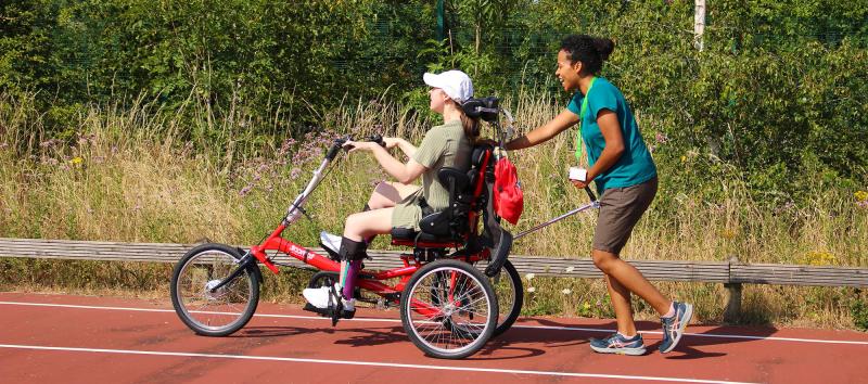 A physiotherapist pushes Treloar's student in an adapted tricycle along a track surrounded by greenery. It is a hot day, both women are wearing shorts and T-shits.