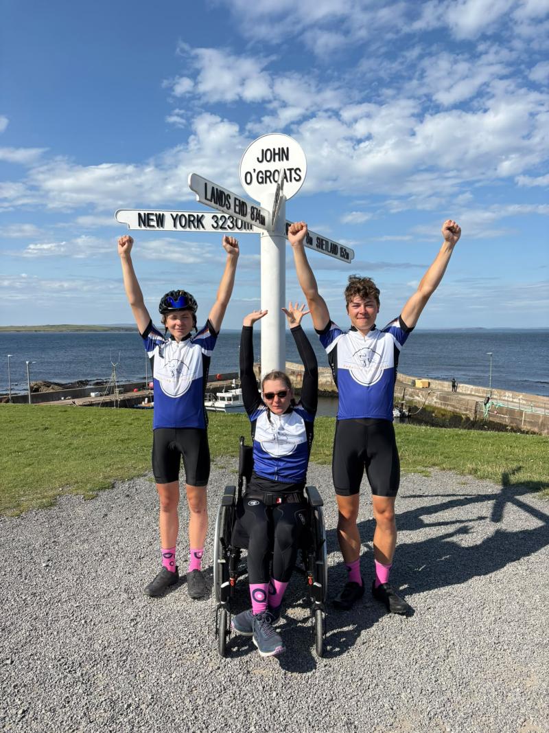 Three cyclists pose in front of John O'Groats signpost, one is in a wheel chair. 