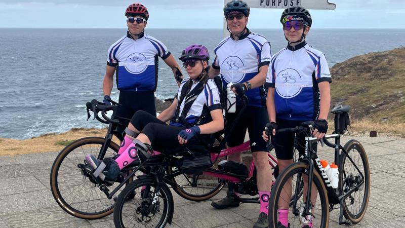 A family of four cyclists stand together in front of a sign post.