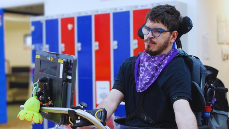Treloar College student using eye gaze to operate his AAC device with colourful lockers in the background. He is wearing glasses and a black T-shirt.