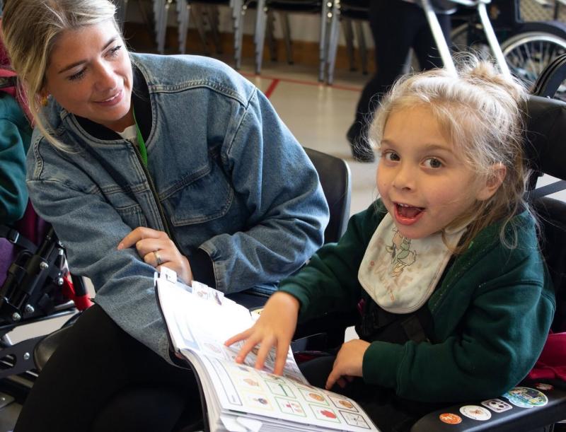 Younger student using a communication book with a support assistant by her side