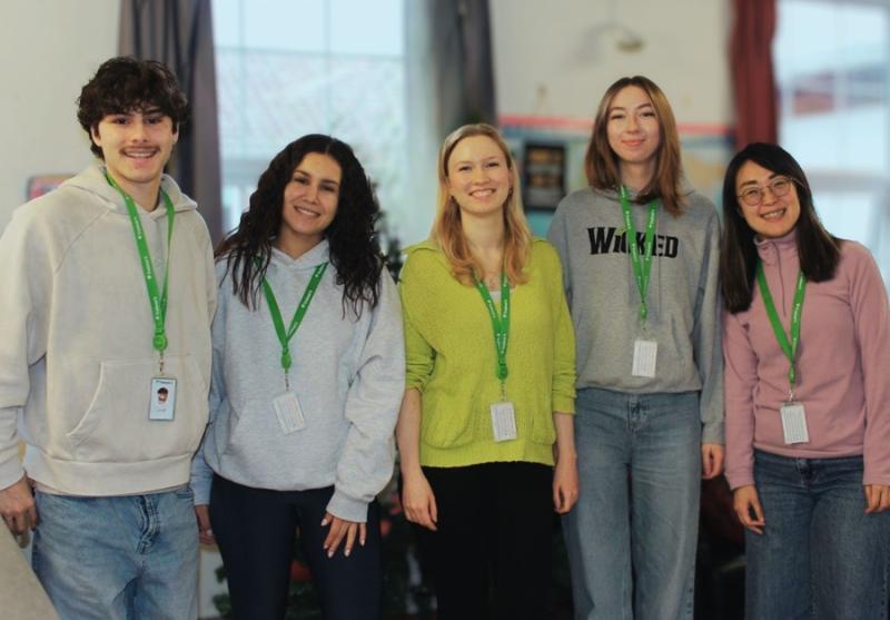 Five young adults wearing ID badges and casual clothes stand smiling in a bright indoor setting.