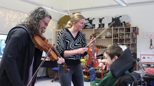 Kate Read, a professional viola player, is playing a viola, standing in front of Treloar's student who is smiling and enjoying the music; Treloar's Head of Music is standing next to them and is holding a violin.