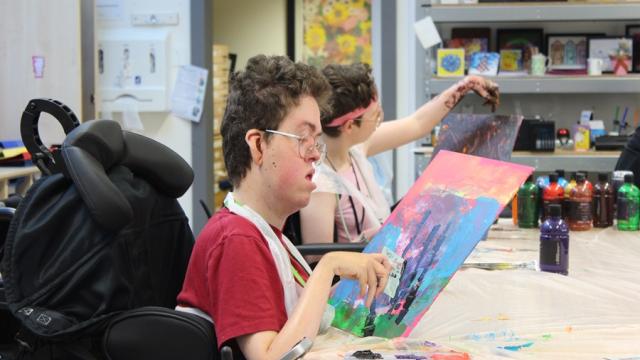 2 Treloar College students at a long desk in the art classroom, working on their painting by using credit cards to place paint. Both are very focused on the task.