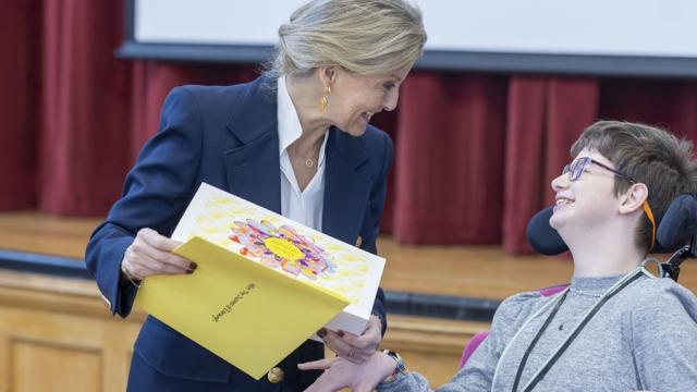 Her Royal Highness The Duchess of Edinburgh is holding a hand-made birthday card created by Treloar College student who is just next to her. Her Royal Highness and the students are both looking at each other and smiling.