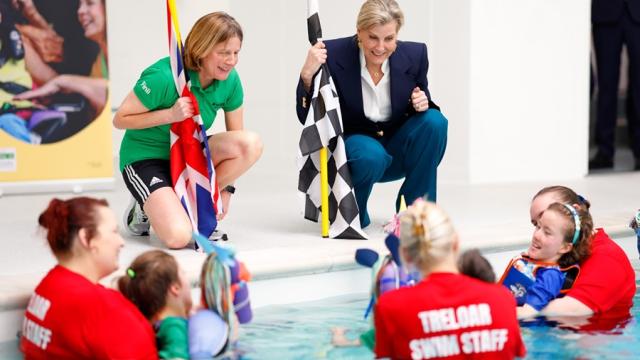 Her Royal Highness The Duchess of Edinburgh talking to Treloar's students who are in the pool with their assistants. The students have just finished a race and Her Royal Highness is holding a chequered flag.