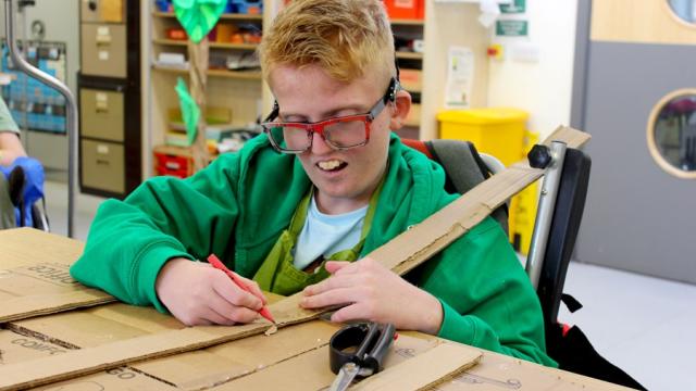 Treloar's student using a pencil to mark where to cut a long piece of cardboard; he is very focused on the task.