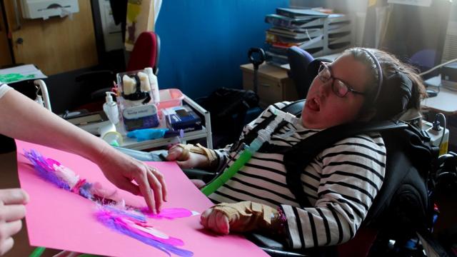  A student support assistant is glueing a pink feather onto a big pink paper after being directed to do so by a Treloar's student who is looking at the stage prop.