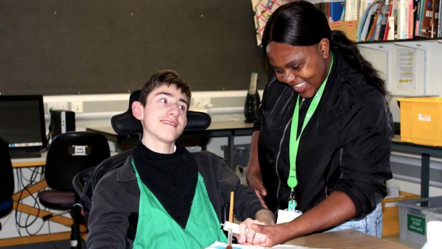 Treloar's student is using a paint brush to paint a stage prop; his assistant is holding his hand to help him paint and hold the brush. They are both smiling.