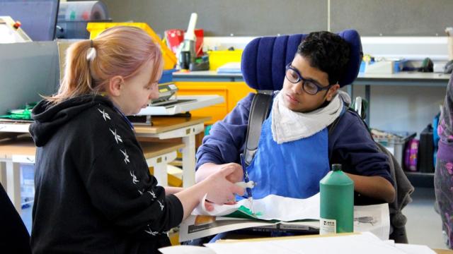 Treloar's student is using a paint brush to paint a stage prop; his assistant is holding his hand to help him paint and hold the brush. they are both really focused on the task.