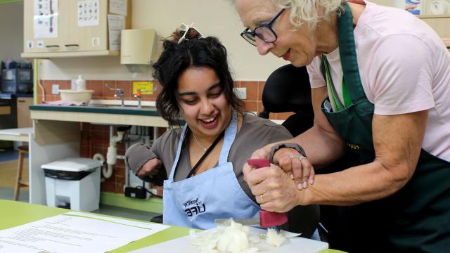 Treloar College student is chopping an onion and her tutor is helping her to do it by holding her hand the the accessible knife.