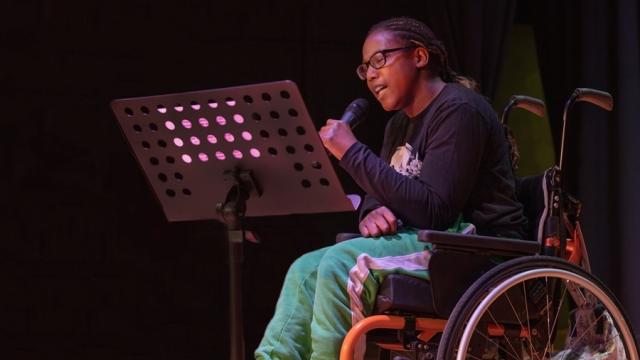 Treloar College student on stage, holding a microphone and singing looking at the lyrics in front of her.