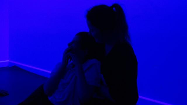 Treloar's student being held by his assistant - both sitting on a mat on the floor during an atmospherics session. The room is lit by a blue light. The student looks mesmerised by this sensory experience.