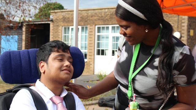Treloar School student is smiling at his student support assistant; she put her hand on his shoulder while talking to him. He is wearing a white shirt with a pink tie.