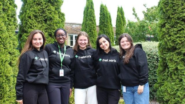 Five female volunteers standing outdoors in front of tall green shrubs, all wearing black hoodies with the 'Treloar's' logo on them.