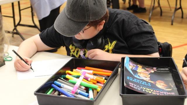Treloar College student wearing a black cap, drawing with a black marker at a table filled with colorful markers and a book. 