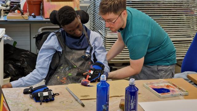 Treloar College wearing a paint-splattered apron is assisted by his assistant in a teal T-shirt in an art classroom filled with supplies and drying racks.