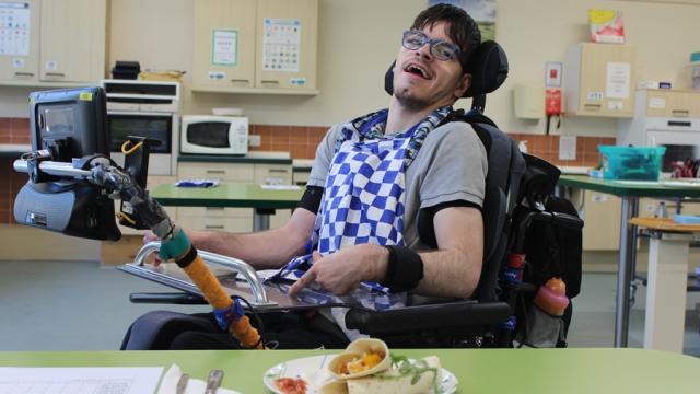 Treloar College student wearing a blue and white checkered apron, at a table with a plate of food he has just cooked including a wrap and salsa. In the background, there are kitchen appliances and tools.