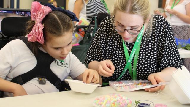 Treloar School student sitting at a table crafting with colorful beads and string, focusing intently on her beadwork activity. Her assistant is sitting next to her assisting her.