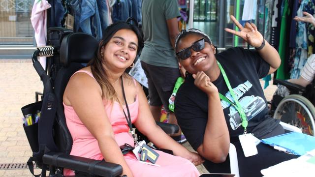 Treloar College student is sitting at a desk with her assistant by her side at an outdoor event.the student is wearing a pink dress, and her assistant is making a peace sign.