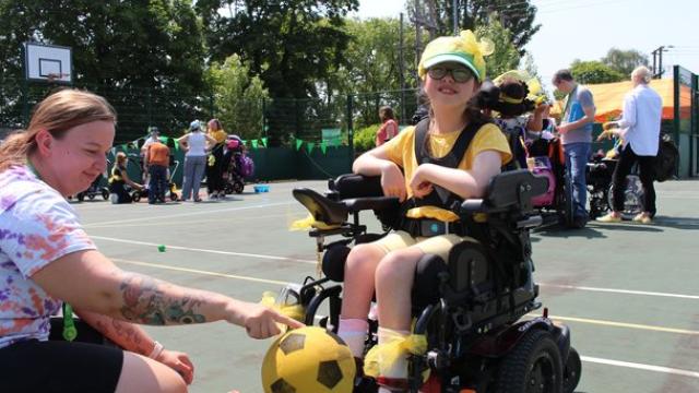 Treloar's primary student wearing yellow accessories plays with a yellow and black soccer ball on an outdoor court during a sunny day during Sports Day. Staff, students and families in the background.
