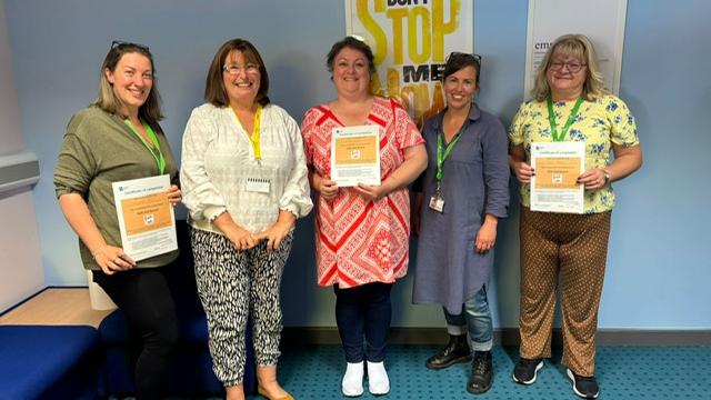 A group of 5 women, lined up in a room, posing for a picture. Some of them are holding certificates. Everyone is smiling.