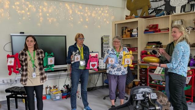 Philippa Perry from BBC Radio 3 with Treloar's staff members during a music lesson: they are all standing in front of the class, holding toys with attached stickers portraying different emotions.