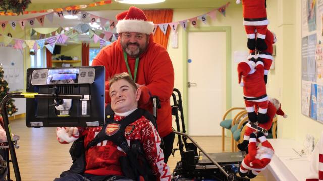 A man in a santa hat is smiling at the camera, with him is a young man in a wheelchair using an AAC device