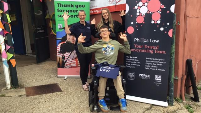 Treloar's student at the Dummer Fair posing for a photo with a sponsor and Treloar's member of staff in front of 2 banners. Everyone is waving at the camera.