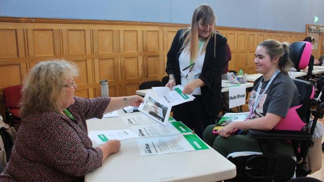 2 Treloar's staff members engage in a discussion with Treloar's student at a table covered with informational brochures inside a wood-paneled room.