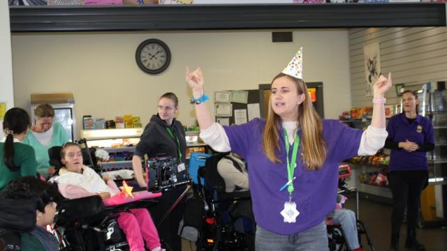 Treloar's Makaton tutor wearing a party hat and purple shirt joyfully raising her arms during a Makaton celebration event with with staff and students around her.