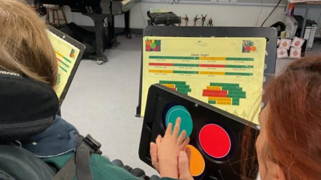 Over the shoulder shot of Treloar's student using a tablet with 3d printed case on which allows the student to play music using Clarion; the student is touching colored buttons on the screen to create music, she is guided by her assistant in a music classroom with a piano and sheet music on stands.