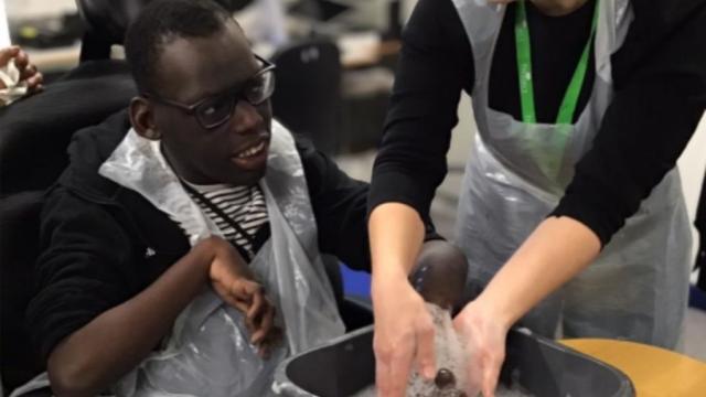 Treloar College student during his session with his occupational therapist: they are in the classroom and the OT is helping him wash his hand in a bowl with water with soap.