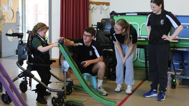 Treloar's primary student in the standing frame playing boccia with college students from a local college assisting her and observing her use a ramp.
