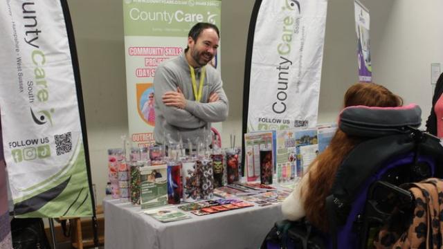 An exhibitor standing behind a CountyCare information booth with brochures and promotional items displayed on the table. He is smiling and listening to Treloar's student in front of him.