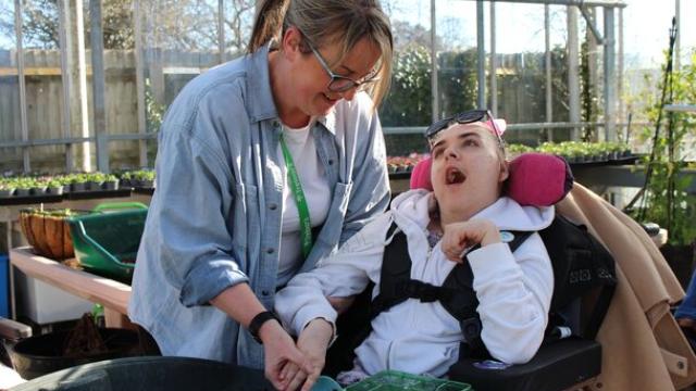 A support assistant assists Treloar's student with planting seeds in a greenhouse filled with gardening supplies.