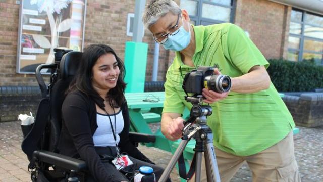 Treloar's student support assistant in green shirt is adjusting a camera on a tripod outdoors for Treloar College student who is about to take some pictures.