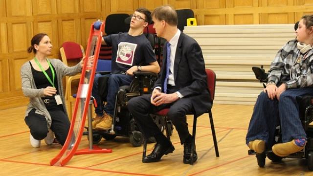 A support assistant kneels beside Treloar's student is demonstrating how to play boccia to Sir Stephen Timms who is wearing a suit and is watching attentively the student.