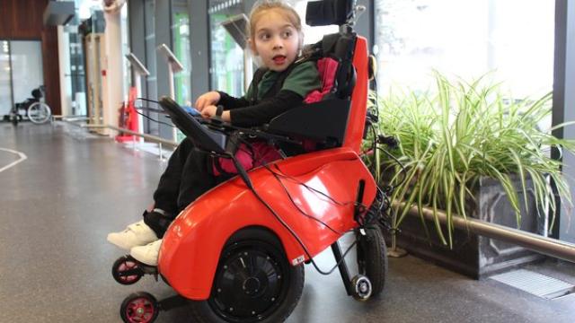 Treloar's primary student seated in a tiny trax chair wheelchair inside a spacious building with large windows and plants nearby.