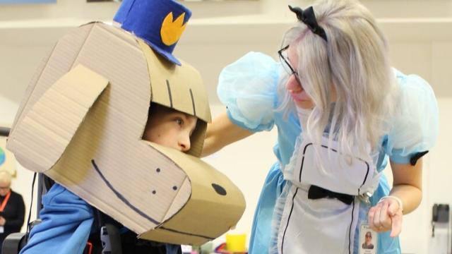Treloar's primary student using a walker, wearing a large cardboard dog head and a hat, interacting with his Student Support Assistant dressed as Alice from Alice in Wonderland in a gymnasium.