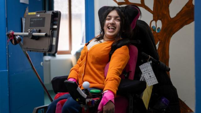 School student in a Powerchair with an AAC Communication device, smiles towards the camera wearing a bright orange cardigan
