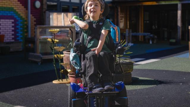 School student in the playground, in the sunshine in a power wheelchair with coloured bricks in a rainbow pattern behind them