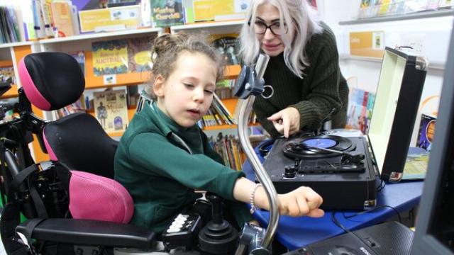 Treloar's primary student interacting with a record player while her assistant guides her in a colourful library setting.