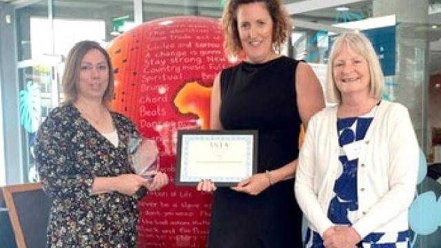 Three women standing indoors, one holding a certificate, with a large red apple-shaped display in the background featuring text. They all look very happy.