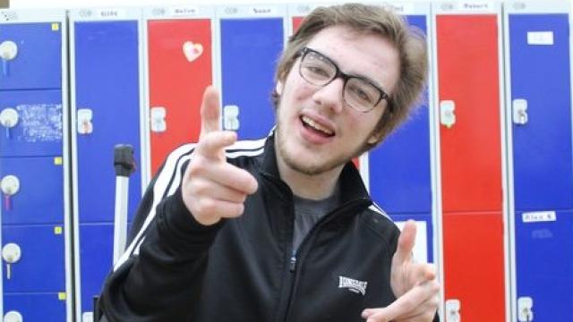 College student in front of a wall of lockers, smiling and pointing at the viewer.