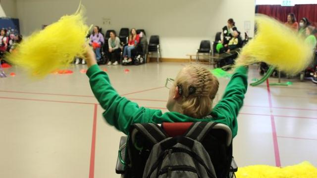 Treloar's student with braided hair enthusiastically waving yellow pom-poms in a gymnasium filled with seated spectators. He is wearing a cochlear implant.