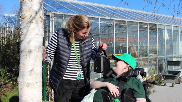 Treloar's teacher in a striped shirt and black vest smiling and interacting with a student outside a large greenhouse on a sunny day. She is holding a speaker near the boys ear so that he can listen to the tree and the sounds of transpiration.