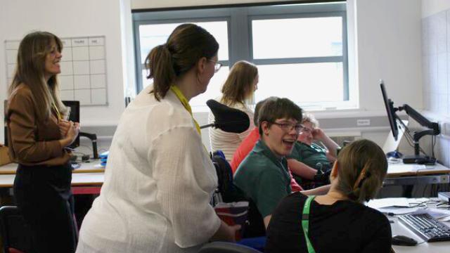Two visitors with  yellow lanyards standing next to Treloar's students who are working on their computers in a classroom. One of the students is smiling and having a lively conversation with the visitor and his assistant.